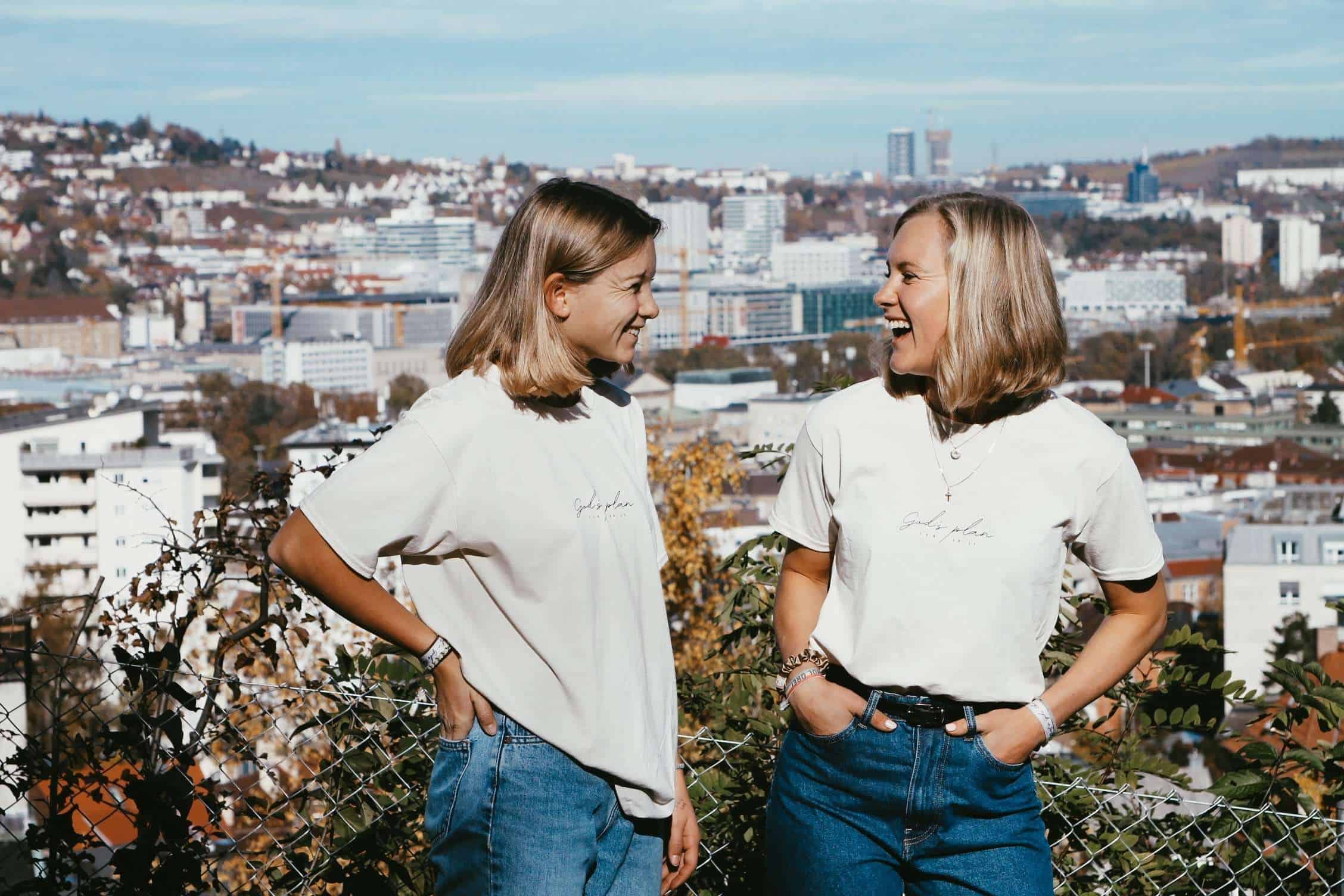 T-Shirt "God's Plan" Unisex 16 Gemeinsames Lachen zweier Frauen mit christlichem T-Shirt, Outdoor, Stadtpanorama, Freude, Freundschaft.