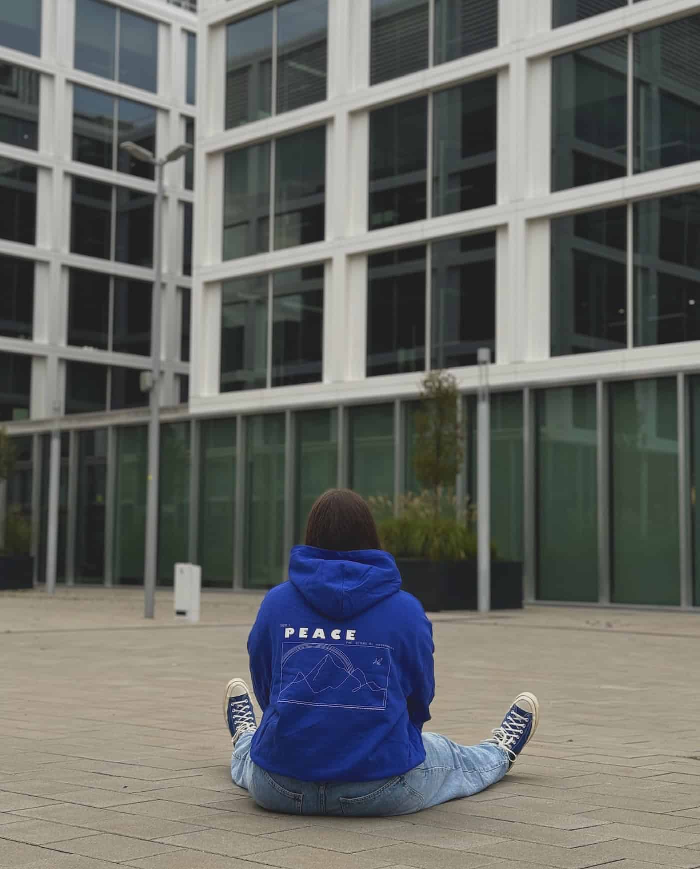 Peace | Christlicher Hoodie – blau unisex – Bio-Baumwolle – Fairtrade 13 Moderne Architektur mit Glasfassade, junge Frau in Hoodi, christliche Marken, spirituelles Bewusstsein, urbaner Lebensstil.