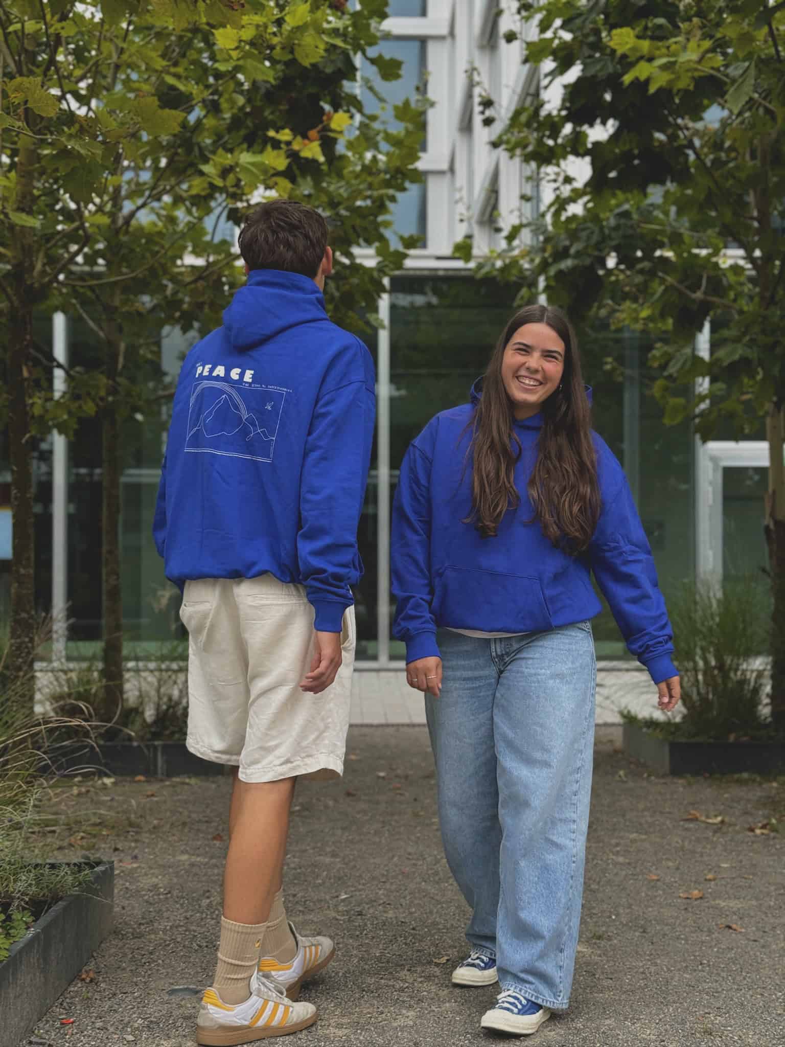 Peace | Christlicher Hoodie – blau unisex – Bio-Baumwolle – Fairtrade 14 Menschen tragen blaue Christliche Hoodies, umgeben von Natur und moderner Architektur.