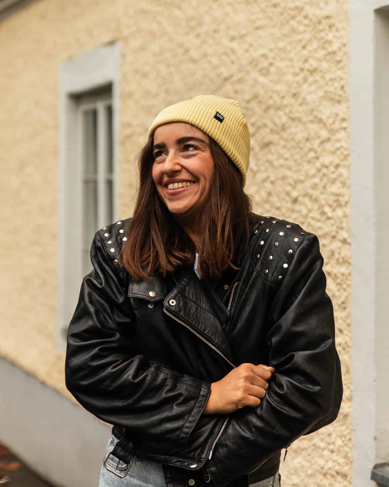 fisherman beanie / gelb / kolosser3 3 Begeisterte junge Frau trägt schwarzen Lederjacke mit Nieten und gelben Strickmütze.