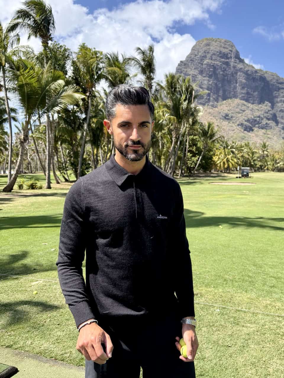 Mann im schwarzen Pullover im Garten mit Palmen und Berg im Hintergrund.
