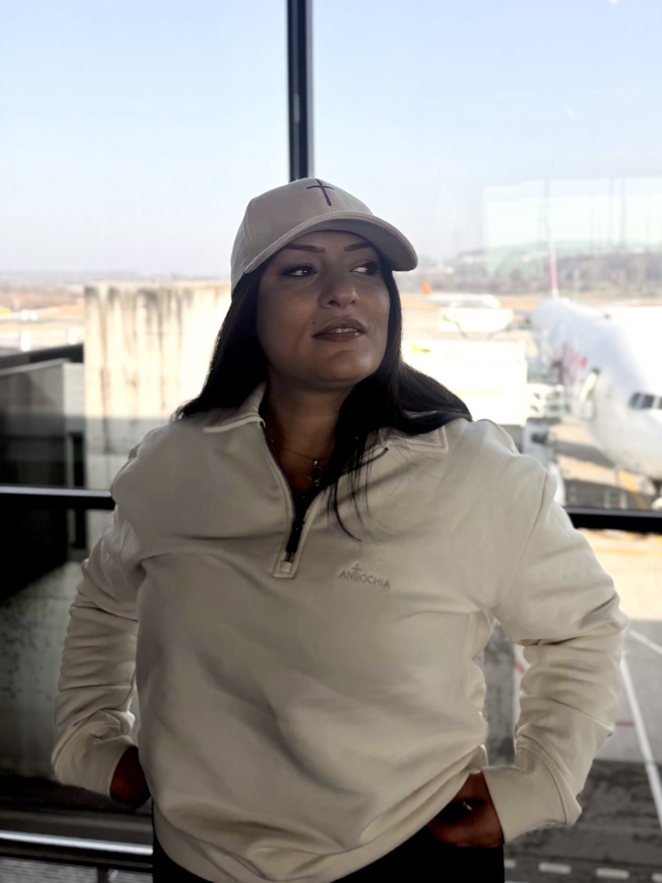 Kord Basecap - "Cross" 18 Gebündelte Frau in Kapuzenjacke und Baseballcap am Flughafen, Flugzeuge im Hintergrund.