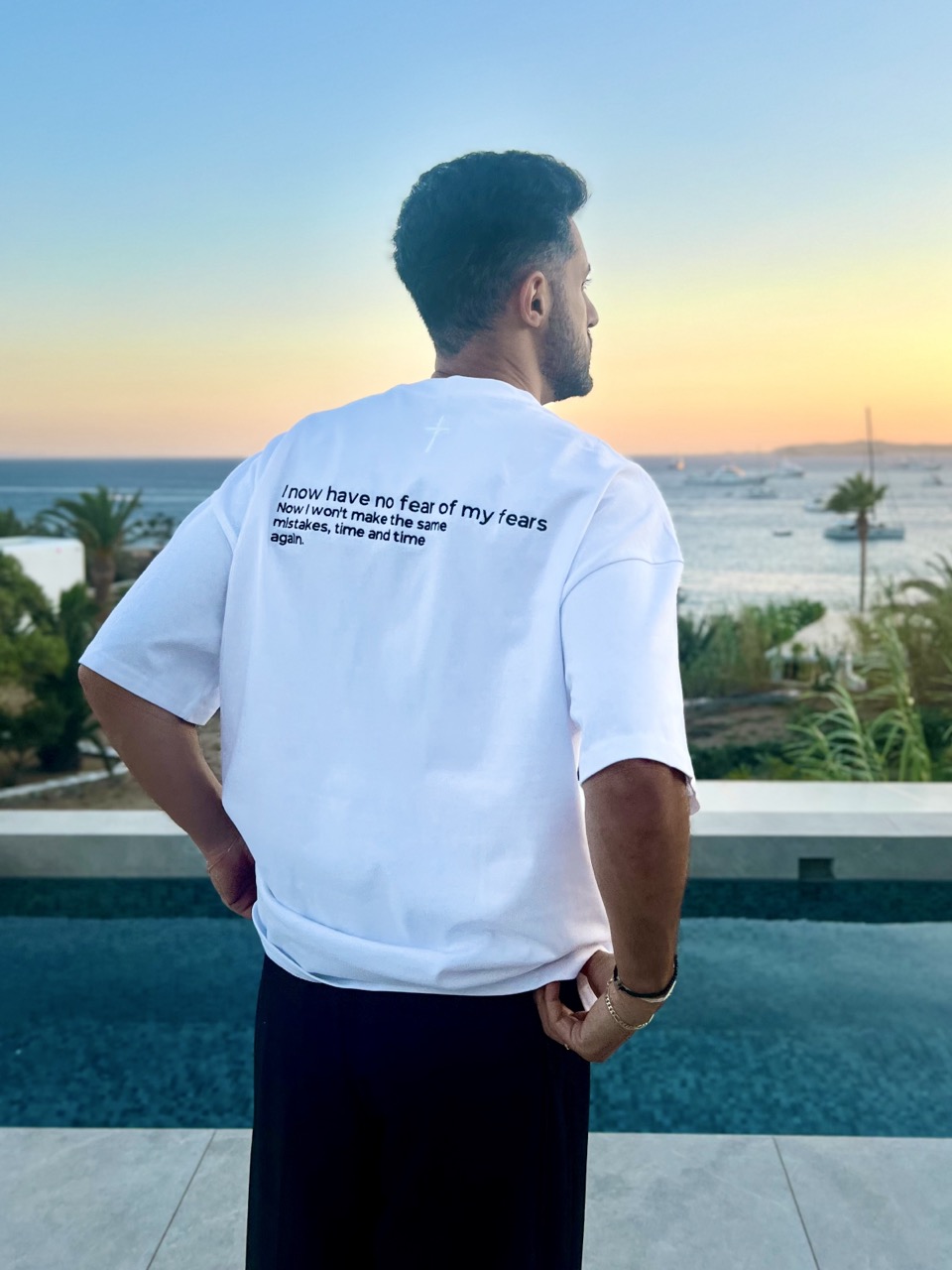 Heavy Oversize T-Shirt - "No Fear" 14 Der Mann blickt auf das Meer bei Sonnenuntergang, trägt ein weißes T-Shirt mit Bibelvers.