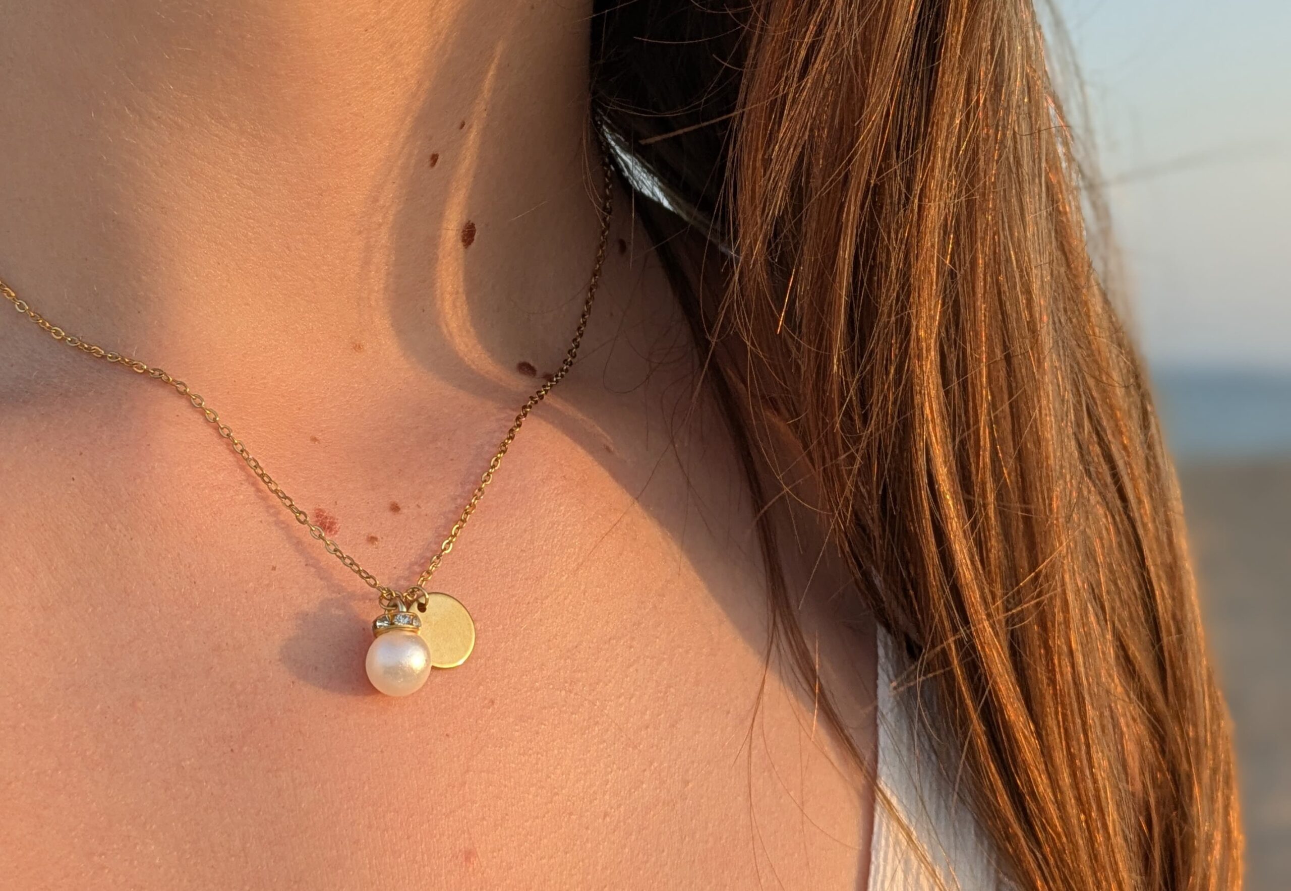 Goldkette "Der dich behütet" 5 Eine Frau trägt eine goldene Halskette mit Perle und Anhänger am Strand bei Sonnenuntergang.