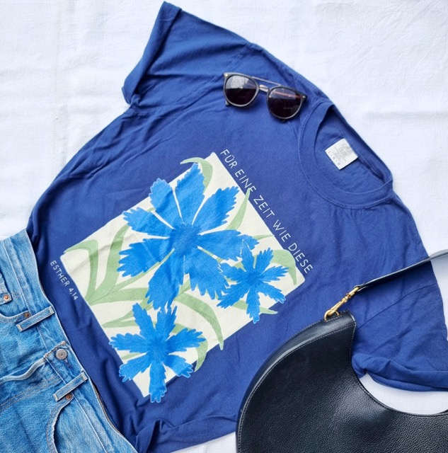 Ein blauer T-Shirt mit Blumenprint und Bibelvers, Sonnenbrille und Tasche auf weißem Stoff.
