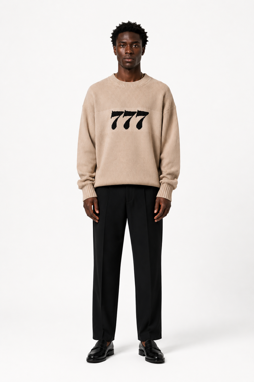Ein moderner Mann trägt einen beigen Pullover mit "777" und schwarze Hosen, christliche Mode.